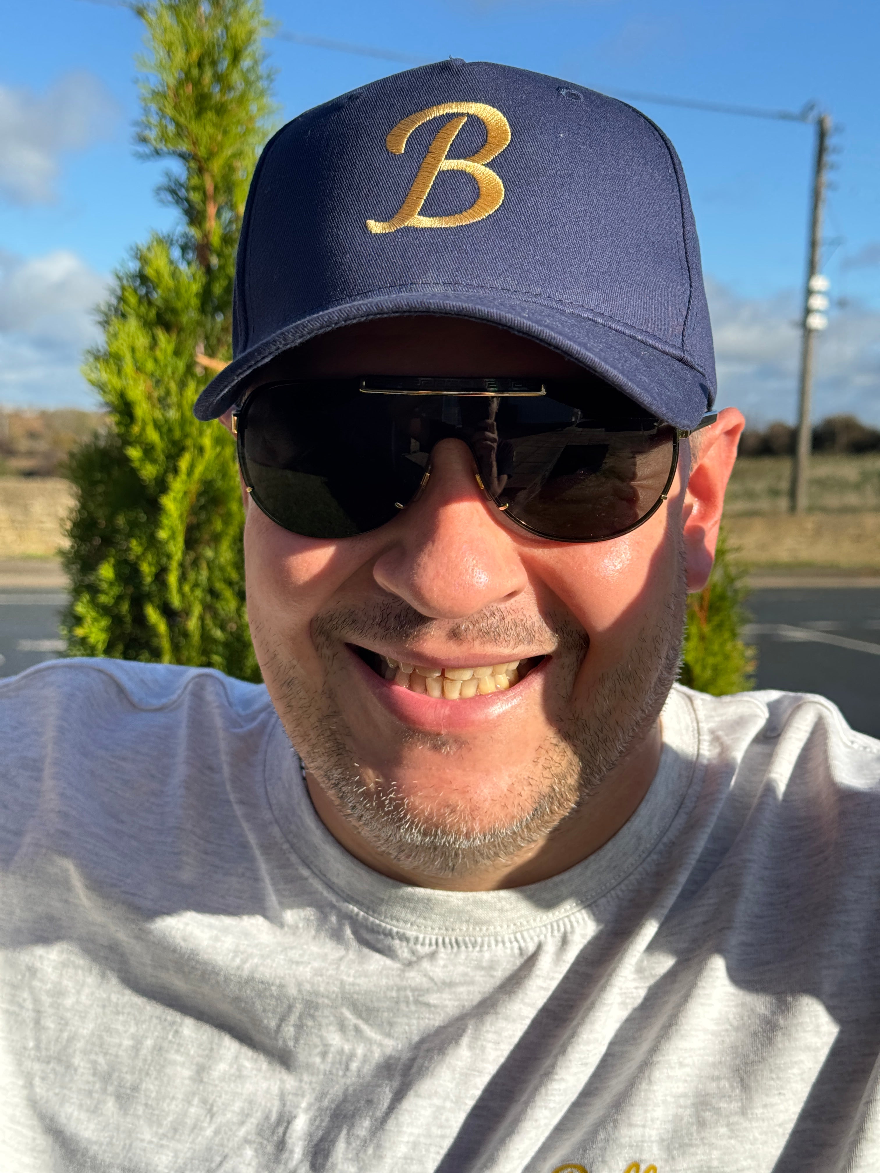 Person wearing a navy cap with a gold 'B' logo and sunglasses, outdoors with trees and blue sky.