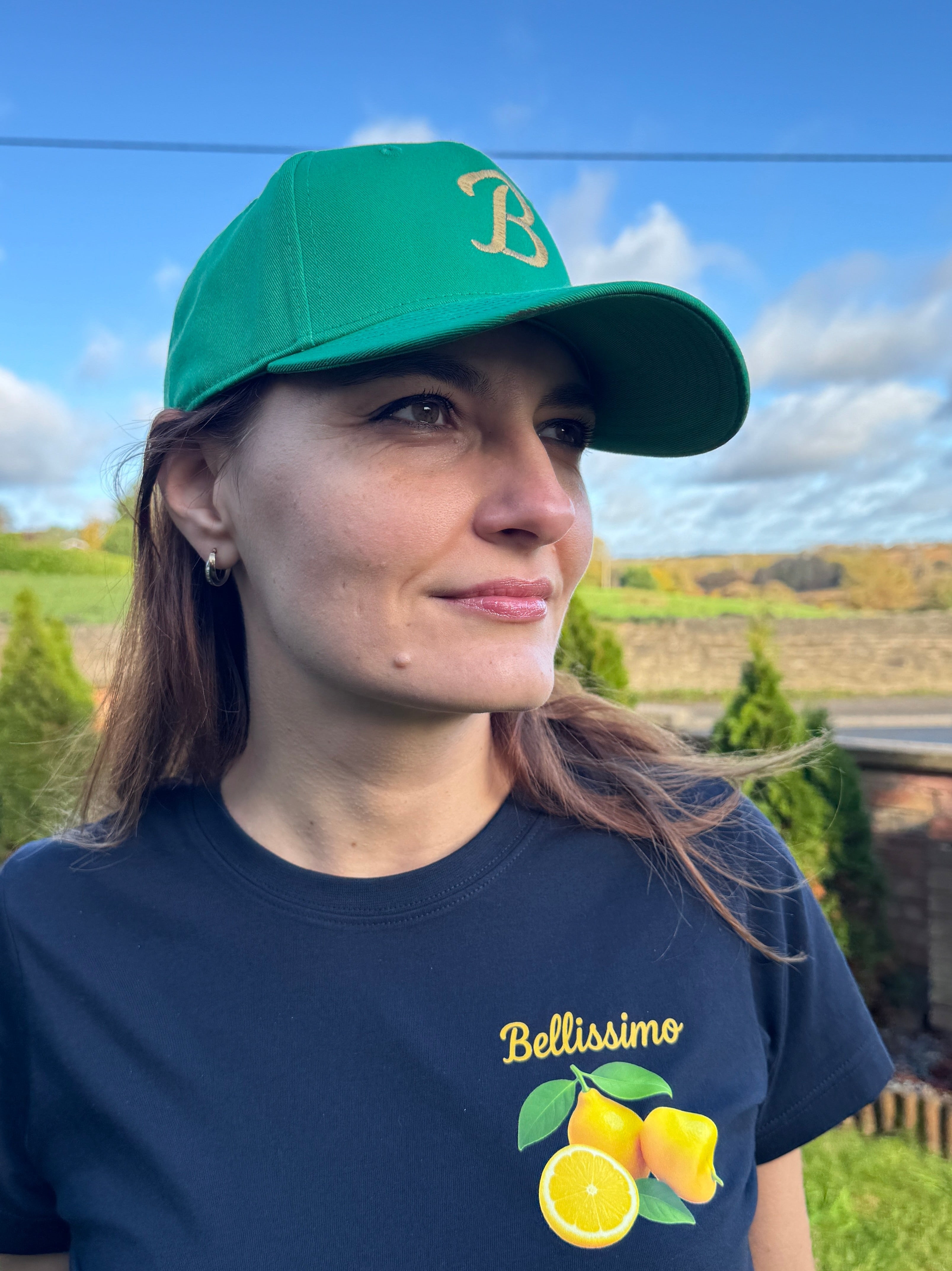 Woman wearing a green cap and navy t-shirt with 'Belissimo' and lemon design outdoors.